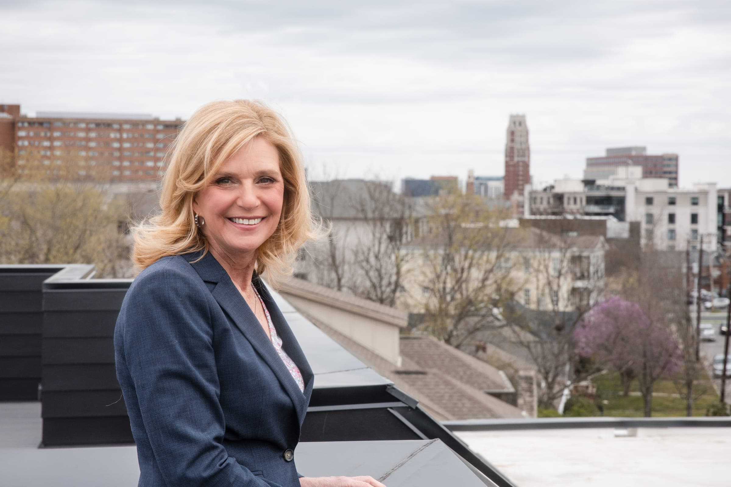 A smiling agent leaning on a rooftop rail with the Nashville skyline and spring treetops spread out behind her